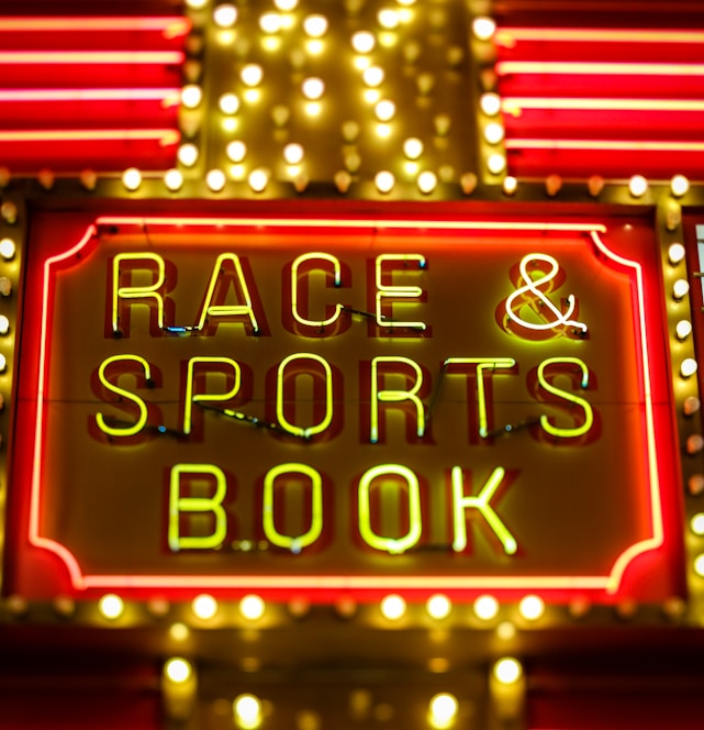 Bright neon lights on a marquee sign display the words 'RACE & SPORTS BOOK'. The sign is surrounded by rows of yellow and white lights against a red background, evoking a vibrant, energetic atmosphere typically associated with casinos or entertainment venues.