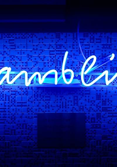 A bright blue neon sign spells out the word 'gambling' in cursive script. The background features a textured wall covered in domino tiles, also illuminated by the blue light, creating a vivid and visually striking scene.