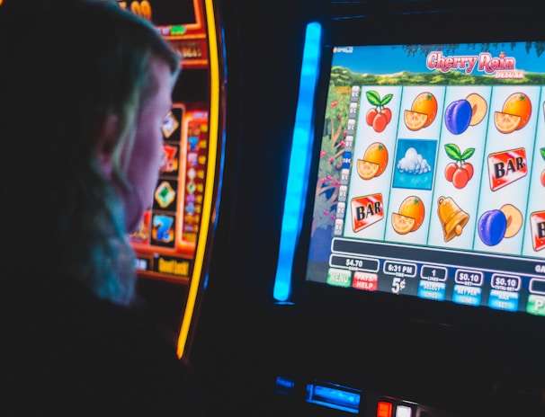 A person is closely observing a digital slot machine with colorful fruit symbols, bars, and a bell. The slot machine screen displays symbols like oranges, plums, cherries, and clouds. The background is dimly lit, creating a focused and somewhat intense atmosphere.